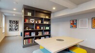 This single-floor amenity space is designed as a social and recreational hub, combining a well-appointed lounge area with a dedicated game room. The open layout is divided into two distinct yet connected zones, separated by a large, dark-toned built-in shelving unit that serves as both a functional game library and a striking architectural focal point. The shelves are stocked with a curated collection of board games, puzzles, and activities, making the space immediately inviting for group gatherings.

The game area centers around a light-colored square table with a built-in lazy Susan, surrounded by modular ottomans in bold yellow and soft teal — a playful color palette that energizes the room. Natural light filters in through a row of windows along one wall, brightening the neutral gray carpeting and white walls. Colorful framed artwork adds personality throughout the space.

The lounge zone features a generous teal sectional sofa dressed with patterned and knotted accent pillows, arranged around a round coffee table. Recessed lighting illuminates both areas evenly, while exposed ceiling beams lend an understated industrial character. A pegboard accent wall, built-in storage cubbies, and a frosted glass entry door complete the space, combining practicality with a modern, thoughtfully designed aesthetic.