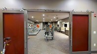 This fitness center offers a thoughtfully designed workout environment spread across a single open floor. The space is accessed through wide sliding barn-style doors framed by rich red accent panels, creating a welcoming and stylish entrance. Inside, the workout floor is covered with patterned carpet that adds a refined touch to the functional space.

The cardio zone lines the left side of the room, featuring a row of treadmills positioned beneath vibrant framed artwork that adds a splash of color to the neutral walls. At the center of the floor, strength training machines are strategically placed to allow for comfortable circulation and safe use. To the right, additional cardio and strength equipment is arranged along the wall, accompanied by a flat-screen television and a dedicated storage or refreshment counter.

The ceiling is fitted with multiple recessed lights providing consistent, even illumination throughout, while ceiling fans ensure proper air circulation during workouts. The warm, neutral color palette of the walls and flooring contributes to a focused yet comfortable atmosphere. Overall, this fitness center balances a professional gym aesthetic with practical amenities, making it a comprehensive and inviting space for a wide range of exercise routines.