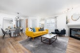 This thoughtfully designed single-floor residence offers a cohesive blend of modern functionality and eclectic style throughout its well-appointed rooms. The open-concept living area is anchored by a fireplace with a tiled surround and light hardwood flooring, creating a warm and inviting atmosphere. Large windows dressed with patterned curtains and white blinds allow generous natural light to fill the space. The living room accommodates comfortable seating and a central coffee table, flowing seamlessly into the dining area, which features a round white table beneath a striking oversized wire pendant light. Adjacent to the dining area, the kitchen is equipped with dark espresso cabinetry, stainless steel appliances including a refrigerator, electric range, microwave, and dishwasher, along with light quartz countertops and a stainless steel sink. Two bedrooms each feature distinctive accent walls — one with a whimsical illustrated wallpaper and another with a bold geometric painted design — along with plush carpeting. The unit includes two full bathrooms: one with a soaking tub and shower combination and another with a sliding glass shower enclosure, both finished with dark wood vanities and white countertops.