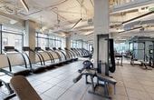 This fitness center is thoughtfully designed to support a full range of workout routines within a spacious, open-plan layout. The facility features exposed ceilings with industrial-style ductwork and bright linear lighting that illuminate the entire floor. Large floor-to-ceiling windows line one wall, flooding the space with natural light while offering views of the surrounding cityscape.

The cardio zone is anchored by an extensive row of treadmills positioned directly along the window wall, maximizing the outdoor views during workouts. The strength training area is well-appointed with a full rack of dumbbells displayed on tiered storage racks in front of wall-to-wall mirrors, along with cable machines, power racks, and adjustable benches to accommodate a wide variety of exercises. Rubber tile flooring throughout provides safety and durability underfoot.

A dedicated secondary strength room offers additional cable machines, a squat rack, and benches in a more intimate setting, also finished with mirrored walls. The facility includes practical amenities such as built-in storage lockers and a towel dispenser station for user convenience. Large glass partitions and sliding doors connect the gym to an adjacent outdoor terrace area, enhancing the sense of openness and providing access to fresh air.