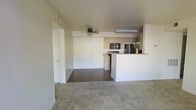 This single-floor space showcases an open-concept layout that seamlessly connects the living area to the kitchen. The living space is finished with neutral-toned carpet, creating a warm and understated foundation. A ceiling fan is centrally positioned overhead, providing both practical air circulation and a subtle design element.

The kitchen is defined by a white peninsula counter that serves as a natural divider between the two areas, offering additional workspace and casual seating potential. Stainless steel appliances, including a refrigerator and microwave, add a modern touch, while the warm-toned wood cabinetry introduces contrast against the predominantly white walls and surfaces. A range and dishwasher complete the kitchen's functional layout. Under-cabinet lighting further enhances the cooking area.

The transition between the carpeted living zone and the hard-surface kitchen flooring clearly delineates the two functional areas while maintaining visual continuity. A doorway on the far left provides access to additional rooms or storage. The overall palette of whites, neutrals, and light wood tones gives the space a clean, cohesive, and move-in ready aesthetic.