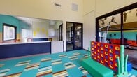 This single-floor children's activity and recreation center is thoughtfully designed to inspire energy and creativity. The main play area is immediately eye-catching, with bold horizontal stripes of teal, yellow, and red adorning the walls, creating a lively and stimulating environment. The space features high ceilings that lend an open, airy feel, while large, dark-framed windows along the exterior walls flood the room with natural light and offer views of the surrounding outdoor greenery.

The play area is well-equipped with oversized building blocks, ride-on toys, and interactive games such as a large Connect Four set, all arranged on a colorful patchwork carpet of striped tiles in teal, multicolor, and neutral tones. Playful wall art, including silhouettes of active figures and decorative cutouts, reinforces the energetic theme throughout.

A standout feature is the enclosed fitness room, separated from the play area by a full-height glass wall with dark metal framing. This transparent partition allows for clear sightlines between the two spaces, making it easy for adults to exercise while supervising children. The fitness area is equipped with exercise machines and receives ample natural light from exterior windows. A reception or check-in counter area is also present near the entrance. Together, these spaces form a comprehensive, family-friendly recreational facility.