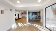 Located at 2850 Uptown Way in Fargo, North Dakota, this single-floor commercial space offers a thoughtfully designed interior that blends modern aesthetics with functional versatility. The entry welcomes visitors through floor-to-ceiling glass double doors framed in dark metal, flooding the foyer with natural light and offering views of the surrounding area.

Inside, the open layout connects several distinct zones. A spacious lounge area near the entrance features a curved sofa, a sculptural pendant light, and framed artwork on the walls, establishing a welcoming and polished first impression. Adjacent to this is a striking blue accent wall adorned with curated art pieces and a wall-mounted display screen, creating a vibrant focal point.

A bar-height counter with acrylic seating and globe pendant lights anchors the social area, backed by an eye-catching mosaic-style blue feature wall that adds texture and depth to the space. A wet bar with dark cabinetry, a subway tile backsplash, and a wall sconce light provides a refined service station nearby.

A semi-private seating nook showcases a vertical wood slat accent wall paired with leather armchairs and a glass-paneled door, offering an intimate retreat within the larger space. Consistent use of light wood-look flooring, recessed lighting, and a neutral white palette unifies the space, giving it a clean, contemporary ambiance throughout.