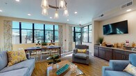The Wireless Lounge in Port Wentworth, Georgia is a thoughtfully designed single-story community space that blends coastal aesthetics with functional amenities. The interior features light wood-tone flooring throughout, warm beige walls, and recessed lighting complemented by decorative pendant and chandelier fixtures with an organic, driftwood-inspired design that reinforces the nautical theme.

The lounge is organized into several distinct zones. A welcoming entry corridor opens into a broad seating area furnished with patterned armchairs arranged around a central coffee table on a navy blue area rug. Beyond this, a main gathering area features a bold cluster chandelier overhead, a glass-top coffee table, plush blue and gray upholstered seating, and coastal artwork on the walls.

Large black-framed windows and glass doors line the perimeter, flooding the space with natural light and offering views of the surrounding community. A dedicated work area includes a herringbone-top wooden table with modern acrylic chairs and a workstation with a monitor, making it well-suited for remote work.

A refreshment station along one wall provides a countertop with a coffee maker and sink, paired with a wall-mounted television above dark cabinetry. Frosted glass sliding barn doors define a private meeting room, adding both style and flexibility to the overall layout.