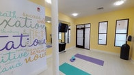 This single-floor yoga and meditation studio offers a calm, purposeful environment designed to support wellness and mindful practice. The space is finished in warm yellow walls that radiate an uplifting, energizing atmosphere, complemented by clean white tile flooring that keeps the overall look fresh and open. A central structural column divides the room subtly without interrupting the generous open floor area, which is outfitted with yoga mats and foam blocks ready for use. One wall features a large, eye-catching typographic mural filled with wellness-inspired words such as "relaxation," "meditation," "namaste," "serenity," and "love," reinforcing the studio's mindful intent. Full-length mirrors are mounted on the walls to assist practitioners in refining their form, while a wall-mounted flat-screen television provides an additional resource for guided sessions. Dark-framed frosted glass partitions and windows line portions of the walls, providing privacy while allowing diffused light to filter through. Flush-mounted ceiling lights ensure consistent, even illumination throughout the space. Storage shelving units offer convenient organization for equipment and accessories. Two interior doors lead to additional areas, and the studio is equipped with climate control for year-round comfort. Overall, the space exudes a serene, focused ambiance ideal for yoga, stretching, and meditation.