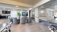 Located at 1585 Kipling St in Lakewood, Colorado, this single-floor community space is thoughtfully designed to foster comfort and social interaction. The open layout flows seamlessly between a well-appointed kitchen and a bright, inviting dining area, all finished with light hardwood-style flooring that unifies the space.

The kitchen is a standout feature, boasting bold slate-blue cabinetry with brushed nickel hardware, striking black-and-white patterned tile backsplash, light quartz countertops, and stainless steel appliances including a full-size refrigerator and dishwasher. Flush-mount lighting and a window provide a clean, functional workspace.

The dining room is bathed in natural light streaming through large sliding glass doors and floor-to-ceiling windows dressed with patterned drapes. Multiple dining tables with colorful geometric-upholstered chairs offer flexible seating arrangements. The doors open directly to an outdoor balcony, extending the social space outside.

A comfortable lounge area rounds out the space, featuring striped barrel chairs arranged around a wall-mounted flat-screen TV, accompanied by a modern multi-ring pendant light and decorative greenery. A wide hallway connects to adjacent corridors, accented by warm wall sconces and handrails, reflecting a thoughtful, accessible design throughout.