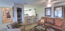 This single-floor residence offers an open and functional layout that seamlessly connects the living area to the kitchen. The living space is anchored by warm, neutral-toned carpeting and features a ceiling fan with a light fixture that contributes to comfortable airflow and ambient lighting throughout the room. A brown leather sofa, accompanied by a patterned armchair and a glass-topped coffee table, creates an inviting seating arrangement centered around a framed wall art piece, adding a touch of refined style to the space.

The kitchen is positioned adjacent to the living area, separated by a raised breakfast bar that accommodates two wrought-iron bar stools, blending practicality with casual dining. White cabinetry lines the upper portion of the kitchen, complemented by green houseplants that introduce natural accents throughout. The kitchen benefits from under-cabinet lighting, enhancing visibility and adding warmth to the cooking area.

To the side of the living space, a built-in shelving unit provides open storage with multiple tiers, ideal for displaying décor or organizing everyday essentials. Overall, the residence presents a cohesive, well-appointed interior with a light, airy palette.