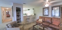 This single-floor residence offers an open and functional layout that seamlessly connects the living area to the kitchen. The living space is anchored by warm, neutral-toned carpeting and features a ceiling fan with a light fixture that contributes to comfortable airflow and ambient lighting throughout the room. A brown leather sofa, accompanied by a patterned armchair and a glass-topped coffee table, creates an inviting seating arrangement centered around a framed wall art piece, adding a touch of refined style to the space.

The kitchen is positioned adjacent to the living area, separated by a raised breakfast bar that accommodates two wrought-iron bar stools, blending practicality with casual dining. White cabinetry lines the upper portion of the kitchen, complemented by green houseplants that introduce natural accents throughout. The kitchen benefits from under-cabinet lighting, enhancing visibility and adding warmth to the cooking area.

To the side of the living space, a built-in shelving unit provides open storage with multiple tiers, ideal for displaying décor or organizing everyday essentials. Overall, the residence presents a cohesive, well-appointed interior with a light, airy palette.