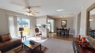 This single-floor space features an open-concept design that seamlessly connects the living room, dining area, and kitchen into one cohesive, functional layout. The living room is carpeted and anchored by a ceiling fan with integrated lighting, a stone-top coffee table, and access to an outdoor patio through a sliding glass door that floods the room with natural light. The kitchen is positioned behind a half-wall breakfast bar, offering partial separation while maintaining an open feel. It is equipped with white appliances, including a full-size refrigerator, and features tile countertops and ample cabinetry. The dining area sits adjacent to the kitchen and is defined by wood-look flooring, a dark wood table with matching chairs, and an elegant pendant light fixture above, complemented by a wall-mounted mirror that adds depth to the space. Neutral-toned walls and a textured ceiling run throughout, creating a consistent, understated aesthetic. The combination of carpet in the living area and hard flooring in the dining and kitchen zones adds both comfort and practicality to this well-organized, light-filled space.