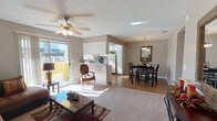 This single-floor space features an open-concept design that seamlessly connects the living room, dining area, and kitchen into one cohesive, functional layout. The living room is carpeted and anchored by a ceiling fan with integrated lighting, a stone-top coffee table, and access to an outdoor patio through a sliding glass door that floods the room with natural light. The kitchen is positioned behind a half-wall breakfast bar, offering partial separation while maintaining an open feel. It is equipped with white appliances, including a full-size refrigerator, and features tile countertops and ample cabinetry. The dining area sits adjacent to the kitchen and is defined by wood-look flooring, a dark wood table with matching chairs, and an elegant pendant light fixture above, complemented by a wall-mounted mirror that adds depth to the space. Neutral-toned walls and a textured ceiling run throughout, creating a consistent, understated aesthetic. The combination of carpet in the living area and hard flooring in the dining and kitchen zones adds both comfort and practicality to this well-organized, light-filled space.