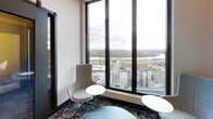 Located on an upper floor of 90 New Street in New Brunswick, NJ, this contemporary study area offers a well-appointed environment designed for focused work and collaboration. The space is organized across a single floor and includes a mix of enclosed study rooms and open lounge-style seating areas.

The private study room features a clean white table with built-in power access ports, surrounded by warm-toned upholstered chairs and a sleek dark sliding barn door. A tall, narrow vertical window admits natural light and frames views of the surrounding cityscape below.

Adjacent to the study room, a bright open lounge area commands expansive floor-to-ceiling windows that offer striking panoramic views of New Brunswick, including the Raritan River and surrounding neighborhoods. This area is furnished with modern high-back lounge chairs and small side tables, creating an inviting setting for individual study or informal meetings.

The corridor connecting the spaces features large-format dark tile flooring, grey walls with wood accent paneling, and black-framed doors with clearly labeled utility and mechanical rooms. The overall design aesthetic throughout is modern and professional, combining neutral tones, contemporary furnishings, and thoughtful architectural details that support a productive academic environment.
