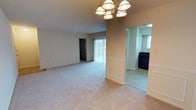 This single-floor space offers a comfortable and functional layout with a warm, understated aesthetic throughout. The main living area is generously proportioned, featuring soft wall-to-wall carpeting in a neutral tone that lends a clean and cohesive look to the space. A classic multi-bulb chandelier serves as a focal point overhead, providing ample ambient lighting and a touch of traditional elegance.

The living area transitions seamlessly into a defined dining space, separated by a wainscoted half-wall with crisp white paneling — a charming architectural detail that adds visual interest and a sense of structure. The wainscoting is echoed along one full wall of the living area, reinforcing a consistent, classic design language throughout.

Natural light filters in through vertical-blind-covered sliding glass doors, offering a view to the exterior and brightening the interior. A doorway on the opposite wall leads to an additional room, providing flexible use of space.

Adjacent to the main living and dining area is a kitchen, visible through an open archway. The kitchen features natural light from its own window and includes a dark-colored dishwasher, suggesting practical, everyday functionality. The overall ambiance of the space is neutral, move-in ready, and well-suited for a variety of interior design styles.