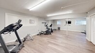 Located at 923 W Main St in New Britain, Connecticut, this dedicated exercise room offers a well-organized fitness environment across a single floor. The space features light hardwood-style flooring throughout, which brightens the interior and provides a clean, modern aesthetic. Bright overhead fluorescent lighting ensures the room is well-lit at all times, supplemented by small windows that allow in natural light.

The fitness area is equipped with a variety of cardio machines, including a treadmill, elliptical trainer, recumbent bike, and a mini trampoline, catering to a range of cardiovascular workout preferences. A dumbbell rack with free weights and an ab bench provide options for strength and core training. A wall-mounted flat-screen TV and a mini-split climate control unit add comfort and convenience to the workout experience.

Full-length mirrors line one wall, enhancing the sense of space and allowing for form checks during exercise. Motivational phrases such as "DEDICATION" and "BEAST MODE" are displayed on the walls, contributing to an energizing atmosphere. A closeted storage area and posted workout charts and rules keep the space organized and functional. Structural support columns are integrated seamlessly into the layout, and the overall design is clean, minimalist, and purpose-built for fitness.
