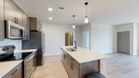 This contemporary single-floor residence showcases a thoughtfully designed open-concept layout with clean lines and modern finishes throughout. The kitchen serves as the centerpiece of the living area, featuring a generously sized island with a white quartz countertop, an undermount sink with a matte black faucet, and stylish pendant lighting overhead. Dark wood-grain cabinetry with black hardware lines the perimeter walls, complemented by stainless steel appliances including a refrigerator, range, and microwave. Light wood-tone flooring flows throughout the main living areas, creating a warm, cohesive aesthetic.

The living room opens directly to a private balcony through sliding glass doors, allowing natural light and outdoor views to fill the space. The carpeted bedrooms offer a comfortable retreat, with the primary bedroom providing access to a walk-in closet and an en-suite bathroom featuring a walk-in shower with subway tile, a single vanity with quartz countertop, and a large framed mirror. A second full bathroom includes a tub-shower combination with the same refined finishes. A dedicated in-unit laundry closet adds everyday convenience to this well-appointed residence.