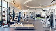 This expansive fitness center is a premier facility designed to accommodate a wide range of workout styles and training needs. The space is defined by its striking architectural details, most notably a large elliptical coffered ceiling with warm ambient uplighting and ceiling fans, lending the main training floor a sense of grandeur and openness. Floor-to-ceiling windows line the perimeter, flooding the interior with natural light and offering panoramic outdoor views that create an energizing atmosphere throughout.

The main floor is thoughtfully divided into distinct training zones. A dedicated cardio area features a full row of treadmills, ellipticals, stationary bikes, and rowing machines, each equipped with individual screens. The free weights section is highlighted by a sculptural white wall with arched, backlit mirrors framing comprehensive dumbbell racks and adjustable benches. Selectorized cable and multi-functional machines are spread throughout, complemented by a functional training zone with TRX suspension systems and a dedicated turf area with a black rig system for battle ropes, medicine balls, speed bags, and plyometric boxes. The durable rubber flooring transitions seamlessly between zones, ensuring both safety and performance across the entire facility.