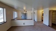 This single-floor residence offers a practical and well-organized open-concept layout designed for comfortable everyday living. The main living area features wall-to-wall carpeting that adds warmth underfoot, while a distinctive half-wall with a wood-toned counter top serves as a subtle divider, defining the space without compromising the open feel. A ceiling fan is centrally positioned overhead, providing both lighting and air circulation to the room. Natural light filters in through a window on the adjacent wall, brightening the interior with an airy feel. The layout flows seamlessly toward the entryway, where a front door is visible alongside additional living space, suggesting a well-connected floor plan. A corridor leads further into the unit, providing access to additional rooms. Glimpses of a kitchen area are visible in the background, featuring light-colored appliances and cabinetry. The neutral wall tones throughout the space create a clean, versatile backdrop well-suited for personalization. The overall ambiance is modest and functional, making efficient use of the available square footage across its single level.