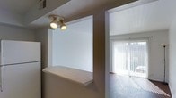 This single-floor space is characterized by a practical, open design that connects the kitchen and living areas through a built-in pass-through counter. The pass-through features a tiled ledge surface, ideal for casual dining or serving, and is illuminated from above by a dual-spotlight ceiling fixture that provides directed task lighting to the area.

The kitchen, partially visible on the left, includes a full-sized refrigerator and benefits from the overhead spotlights. The pass-through opening creates a sense of openness between spaces while maintaining a defined separation between the cooking and living zones.

The adjacent living area is bright and airy, with large floor-to-ceiling windows dressed in vertical blinds that allow abundant natural light to filter through. The hard-surface flooring in the living area reflects the incoming daylight, enhancing the overall brightness of the space. Neutral-toned walls throughout contribute to a clean, understated aesthetic. The corridor connecting both spaces is wide and unobstructed, allowing for smooth flow between rooms. Overall, the space offers a functional and light-filled environment with a straightforward, no-frills design approach.