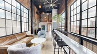 Located in Cincinnati's vibrant urban core, this two-floor space masterfully blends industrial heritage with contemporary design. The ground floor opens into a spacious lounge and gathering area defined by soaring ceilings, exposed brick walls, and dramatic floor-to-ceiling factory-style windows that flood the interior with natural light and frame sweeping city views. Polished concrete floors, large ceiling fans, and pendant lighting contribute to the raw yet refined aesthetic. A bar-height counter and seating arrangements make this level well-suited for both collaborative work and casual socializing. A striking staircase with blue-painted treads and sleek metal railings connects the two floors, serving as an architectural focal point in its own right. The upper floor continues the industrial character with more exposed brick and black-framed windows revealing downtown Cincinnati landmarks. The compact kitchen on this level features clean white cabinetry, a stainless steel undermount sink, and subway tile backsplash, with city views framed directly above the countertop. The bathroom is thoughtfully appointed with penny-tile flooring, a wall-mounted sink, accessibility grab bars, and a decorative geometric gold wall installation. Hallways on both levels ensure smooth circulation throughout the space.