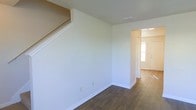 This two-story space presents a straightforward, functional design characterized by clean white walls and dark hardwood-style flooring that runs throughout the main level. The main living area is open and adaptable, with a staircase positioned along one wall providing access to the upper floor. The crisp white walls and white trim create a bright, neutral backdrop that lends itself well to a variety of interior styles. A doorway leads toward the entryway, where a window beside the front door allows natural light to filter into the space, brightening the transitional area between the entrance and the living area. The ceiling features a standard height with visible HVAC venting, ensuring climate comfort throughout. The overall design is minimal and move-in ready, offering a clean canvas for personalization. The consistent flooring and neutral color palette across the visible areas contribute to a cohesive and organized feel throughout the main level.