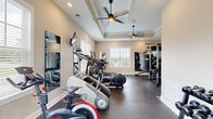 This dedicated fitness center is a comprehensively outfitted single-floor space designed to support a full range of workout routines. The room features elegant tray ceilings with crown molding, recessed lighting, and dual ceiling fans that keep the space comfortable during intense training sessions. Speckled rubber flooring covers the main workout area, providing durability and impact absorption, while a section near the entrance transitions to hardwood-style flooring with interlocking foam mats.

The cardio zone is well-stocked with multiple treadmills, elliptical trainers, a StairMaster, and a Spirit cycling bike, offering a variety of aerobic training options. A wall-mounted flat-screen television is positioned for viewing during cardio sessions. The strength training area includes a multi-station cable machine, a dedicated weight bench, an Everlast heavy punching bag, kettlebells, medicine balls, and a full rack of dumbbells — accommodating everything from functional training to powerlifting.

Full-length mirrors are strategically placed throughout the space, enhancing the sense of openness and allowing for form checks during exercise. Natural light streams in through multiple windows with plantation shutters, offering views of the surrounding greenery. A compact refrigerator near the entrance provides convenient hydration access, rounding out a thoughtfully designed, professional-grade fitness environment.