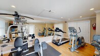 Located in Madison, Alabama, this single-floor fitness center is thoughtfully designed to provide a complete workout environment within a welcoming, neatly finished space. The room features warm-toned walls, crisp white crown molding, and smooth hardwood-style flooring that lend a clean, polished aesthetic throughout.

The space is equipped with a variety of cardio and strength training equipment, including multiple treadmills, an elliptical trainer, a stationary bike, and a multi-station cable weight machine. A large wall-mounted mirror runs along one side of the room, visually expanding the space and allowing users to monitor their form during exercise. Recessed lighting overhead, combined with natural light from multiple windows with blinds, keeps the environment bright and well-lit.

A wall-mounted flat-screen television offers entertainment during workouts, while a standing water cooler ensures easy access to hydration. The space also includes a wall-mounted air conditioning and heating unit for year-round comfort. A set of French-style glass doors at one end of the room provides additional natural light and convenient access. Storage space is available via a set of double closet doors. Safety is addressed with a prominently mounted fire extinguisher. Overall, this fitness center delivers a functional and comfortable environment well-suited for a range of exercise routines.