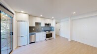 This single-floor space showcases a clean, modern design characterized by crisp white walls, light wood flooring, and recessed lighting throughout. The kitchen is thoughtfully outfitted with white cabinetry extending to the ceiling, a textured tile backsplash, a stainless steel gas range, built-in microwave, dishwasher, and a full-size refrigerator — providing a complete and functional cooking environment. The open layout adjacent to the kitchen allows for flexible use of the living area, which is illuminated by recessed lighting and features a wall-mounted mini-split air conditioning unit for climate control. Sliding glass doors open directly onto a private balcony with a paved tile floor and a metal grid railing, offering a pleasant view of mature trees and greenery that bring a natural, serene quality to the space. The bathroom is finished with attention to detail, featuring tile-surround walls, a glass sliding door enclosing a shower-tub combination, a stone-look countertop vanity with an undermount sink, modern chrome fixtures, and a large mirror. A frosted glass sliding door provides access to a secondary space, ensuring privacy throughout. Natural light filters in through frosted and clear windows, contributing to the overall bright and airy ambiance of this well-designed unit.