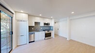 This single-floor space showcases a clean, modern design characterized by crisp white walls, light wood flooring, and recessed lighting throughout. The kitchen is thoughtfully outfitted with white cabinetry extending to the ceiling, a textured tile backsplash, a stainless steel gas range, built-in microwave, dishwasher, and a full-size refrigerator — providing a complete and functional cooking environment. The open layout adjacent to the kitchen allows for flexible use of the living area, which is illuminated by recessed lighting and features a wall-mounted mini-split air conditioning unit for climate control. Sliding glass doors open directly onto a private balcony with a paved tile floor and a metal grid railing, offering a pleasant view of mature trees and greenery that bring a natural, serene quality to the space. The bathroom is finished with attention to detail, featuring tile-surround walls, a glass sliding door enclosing a shower-tub combination, a stone-look countertop vanity with an undermount sink, modern chrome fixtures, and a large mirror. A frosted glass sliding door provides access to a secondary space, ensuring privacy throughout. Natural light filters in through frosted and clear windows, contributing to the overall bright and airy ambiance of this well-designed unit.