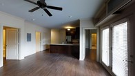 This single-story space showcases an open-concept layout that seamlessly connects the living area to the kitchen, creating a cohesive and functional environment. Dark hardwood flooring runs throughout, providing a rich, warm foundation that contrasts with the light-toned walls and ceilings.

The kitchen is a standout feature, equipped with dark cabinetry that extends to the upper walls, a stainless steel refrigerator with an integrated ice and water dispenser, and a peninsula-style island with a dark wood base and a lighter countertop. The island serves as both a workspace and a natural dividing element between the kitchen and living areas.

A ceiling fan with dark blades is centrally positioned in the living area, contributing to both airflow and aesthetic detail. Recessed lighting and a pendant fixture over the kitchen provide layered illumination throughout the space.

Large windows along one wall allow natural light to filter in, brightening the interior. Additional doorways lead to what appears to be a bathroom and further rooms, suggesting a practical and well-organized floor plan. The overall ambiance is modern and understated, with a consistent dark-toned design palette.