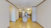 This single-floor space showcases an open-concept layout that seamlessly connects the living area to the kitchen, creating a bright and functional environment. Light hardwood flooring extends throughout, lending warmth and continuity to the space.

The kitchen is a standout feature, equipped with white cabinetry accented by glass-front upper cabinets and a decorative tile backsplash. Stainless steel appliances, including a full-size refrigerator and range, complement the clean aesthetic. A central island with a granite countertop and an integrated sink serves as both a prep area and a casual gathering spot, illuminated by elegant pendant lights overhead.

Crown molding runs along the ceiling line, adding a refined architectural detail to the otherwise modern design. Structural columns with matching trim integrate naturally into the open floor plan without interrupting the flow of the space. Recessed and track lighting supplement the pendant fixtures, ensuring the area is well-lit throughout.

The entry door, finished in a warm gray tone, is visible from the main living area, reinforcing the open sightlines that define this space's welcoming and practical character.