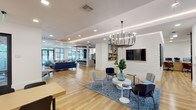 Located at 5840 Farrington Rd in Chapel Hill, NC, this single-floor amenity space offers a thoughtfully designed, open-concept layout that seamlessly blends comfort, functionality, and contemporary style. Light-toned hardwood flooring runs throughout, unifying the various distinct zones and lending a warm, cohesive feel to the entire space.

The main lounge area is anchored by plush navy blue sofas, curated seating arrangements, and a striking multi-arm chandelier that draws the eye upward toward the tray ceilings with integrated ambient lighting. Large black-framed windows allow generous natural light to flow in and provide views of the lush outdoor surroundings. A white brick accent wall hosts a mounted flat-screen TV, while decorative greenery adds life and color throughout.

A dedicated entertainment lounge features a wood-planked accent wall with dual mounted screens, leather armchairs, and a bold landscape mural, creating an inviting, relaxed atmosphere. A separate seating nook with a sleek linear fireplace set into a clean white surround offers an intimate retreat, framed by dramatic blue-tinted glass partition walls.

The kitchenette area is well-appointed with stainless steel appliances, a quartz countertop, under-counter refrigerators, and stylish pendant lighting overhead exposed wooden ceiling beams. A chalkboard-style feature wall anchors a casual dining area nearby, completing this versatile and elegant community space.