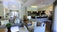 This single-floor space showcases a thoughtfully designed open-concept layout that seamlessly connects the living and kitchen areas into one cohesive, inviting environment. The living area is anchored by plush gray sofas arranged around a wood-and-metal coffee table atop a soft area rug, creating a comfortable and welcoming atmosphere. Decorative crown molding, recessed lighting, and a soaring ceiling with tray details add architectural elegance throughout the space.

The kitchen is a standout feature, boasting rich dark cabinetry with stone tile backsplash, sleek pendant lighting, and a spacious island with bar-height seating for multiple guests — ideal for both casual dining and entertaining. Stainless steel appliances complete the modern culinary setup. Decorative columns with classical detailing help define the transition between the living and kitchen zones while preserving the open feel of the floor plan.

Natural light filters in through large windows and glass-paned entry doors, brightening the neutral warm-toned walls and dark hardwood flooring. The overall ambiance strikes a balance between sophisticated and comfortable, with upscale design elements that create a polished, welcoming environment suited for both relaxation and social gatherings.
