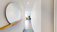 This contemporary single-floor unit is thoughtfully designed with a clean, modern aesthetic and an efficient open layout. A welcoming entryway corridor, accented by a round mirror and warm wood shelf, leads into the heart of the home. The open-plan living and dining area is bathed in natural light from expansive floor-to-ceiling windows that frame striking panoramic city views, creating a bright and airy atmosphere. The living space comfortably accommodates a sectional sofa and accent seating, complemented by hardwood-style flooring throughout. The kitchen is a functional centerpiece, featuring a waterfall island with bar seating, white cabinetry, wood-tone upper cabinets, stainless steel appliances including a refrigerator and microwave, and a chevron tile backsplash. A dedicated in-unit laundry closet adds everyday convenience. The bedroom continues the floor-to-ceiling window theme, offering impressive elevated city skyline views and ample space for a full-size bed and furnishings. The bathroom is finished with penny-tile flooring, a wood-vanity with white countertop, and a mirrored medicine cabinet, delivering a polished, spa-like feel. Recessed lighting and a neutral white palette unify the space with a crisp, contemporary character.