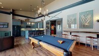 Located at 5412 Edwards Farms Rd in Columbus, Ohio, this single-floor recreational space combines rustic lodge aesthetics with refined design details throughout. The main gathering area centers on a full-size pool table with blue felt, anchored beneath a dramatic rope-and-antler chandelier and soaring ceilings accented by bold dark crown molding. Vintage ski décor and wall art add character to the elevated upper walls, reinforcing the space's distinctive lodge-inspired theme. Rich hardwood flooring flows seamlessly throughout, tying each area together cohesively. The kitchenette is outfitted with custom slate-blue cabinetry, dark granite countertops, a tile backsplash, and full appliances including a refrigerator and microwave. Rustic twig-style bar stools line the kitchen island, and an antler chandelier above echoes the space's cohesive design language. A dedicated lounge area features large multi-pane windows dressed in bold red patterned drapes, offering generous natural light and views of an outdoor pool. Leather seating and a reclaimed wood coffee table furnish this bright corner, creating an inviting retreat. Additional high-top seating near the billiards area and wall-mounted cue storage complete the functional, entertaining-focused layout. Wall sconces and recessed lighting ensure warm, layered illumination throughout.