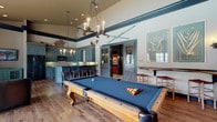 Located at 5412 Edwards Farms Rd in Columbus, Ohio, this single-floor recreational space combines rustic lodge aesthetics with refined design details throughout. The main gathering area centers on a full-size pool table with blue felt, anchored beneath a dramatic rope-and-antler chandelier and soaring ceilings accented by bold dark crown molding. Vintage ski décor and wall art add character to the elevated upper walls, reinforcing the space's distinctive lodge-inspired theme. Rich hardwood flooring flows seamlessly throughout, tying each area together cohesively. The kitchenette is outfitted with custom slate-blue cabinetry, dark granite countertops, a tile backsplash, and full appliances including a refrigerator and microwave. Rustic twig-style bar stools line the kitchen island, and an antler chandelier above echoes the space's cohesive design language. A dedicated lounge area features large multi-pane windows dressed in bold red patterned drapes, offering generous natural light and views of an outdoor pool. Leather seating and a reclaimed wood coffee table furnish this bright corner, creating an inviting retreat. Additional high-top seating near the billiards area and wall-mounted cue storage complete the functional, entertaining-focused layout. Wall sconces and recessed lighting ensure warm, layered illumination throughout.