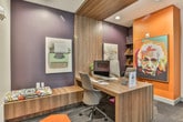Located in Atlanta, GA, this single-floor business center combines functional design with a distinctly creative aesthetic. The space is anchored by a reception area featuring a sleek wood-finished desk, recessed lighting, and a bold pop-art portrait of Albert Einstein set against a striking orange accent wall — immediately establishing the space's energetic character.

The workspace area offers dedicated computer workstations equipped with iMac desktops, arranged along a continuous wood-grain desk surface. Dark charcoal walls contrast with white trim and contemporary framed artwork, including vintage-themed prints of a typewriter and rotary phone, lending a playful, intellectual ambiance. Ergonomic seating and built-in low-profile storage benches with colorful accent cushions provide both comfort and practicality.

Throughout the space, patterned carpet tiles in bold orange, purple, and gray create visual energy underfoot, while geometric hexagonal area rugs add further design interest. A corridor connects the workstations to restroom facilities and additional rooms enclosed by black-framed glass doors, ensuring a sense of openness while maintaining defined zones. Recessed ceiling lights illuminate the entire space consistently, creating a professional yet stimulating environment well-suited for focused work.