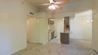 This single-floor space features an open-concept layout that integrates the living area and kitchen into one cohesive environment. The living area is finished with neutral-toned carpet and painted walls, creating a clean, understated aesthetic. A ceiling fan with a built-in light fixture is centrally mounted overhead, providing both airflow and ambient lighting throughout the space.

The kitchen is positioned adjacent to the living area and is defined by a raised peninsula with a laminate countertop, offering a convenient surface for casual dining or meal preparation. Cabinetry in a warm wood-tone finish lines the upper kitchen walls, complementing the white appliances below, which include a refrigerator, dishwasher, and range. A small television is mounted within the kitchen cabinetry, adding a functional touch to the cooking space.

A hallway extends from the living area, suggesting additional rooms beyond. A set of closet doors is visible along one wall, providing ample storage. The overall color palette is light and neutral throughout, contributing to a bright and airy atmosphere. The layout is practical and efficient, making effective use of the available space.