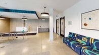 Located at 10401 Motor City Dr in Bethesda, MD, this thoughtfully designed pet amenity space offers a comprehensive environment for dogs and their owners. The open-plan layout is anchored by polished concrete flooring and white structural columns, creating a clean, modern aesthetic throughout. Overhead, cylindrical pendant light fixtures provide consistent, well-distributed illumination across the entire space.

The play area occupies one end of the space, featuring carpeted flooring and an array of dog agility and play equipment, including ramps, tunnels, and a hoop obstacle. Large windows along this section flood the area with natural light and offer views of the surrounding greenery, while a wall-mounted bone-shaped sign reading "home is where your dog is" adds a playful touch. Colorful, boldly patterned seating runs along the perimeter, providing comfortable spots for owners.

The grooming zone is equipped with multiple stainless steel elevated wash basins fitted with handheld sprayers, alongside grooming tables on adjustable stands. The walls in this area feature a textured concrete finish, lending an industrial refinement. A teal graphic frieze runs along the upper portion of one wall, adding a vibrant design element that ties the space together. The overall ambiance is energetic, functional, and pet-forward.