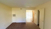 This two-floor residence offers a practical and functional layout designed to maximize usability across its levels. The main living area features wall-to-wall carpeting that adds warmth underfoot, while neutral-toned walls and a textured ceiling create a clean, understated aesthetic throughout. A built-in wall unit provides integrated ventilation, contributing to the room's efficient design. Natural light filters generously into the space through vertical blinds that line a bright sliding door or window opening, illuminating the adjoining area and enhancing the overall sense of openness. The transition between rooms is seamless, with doorways connecting the carpeted living space to an adjacent area with smoother flooring, suggesting a natural separation between living and utility zones. Ceiling-mounted HVAC vents are present throughout, ensuring consistent climate control across the space. The staircase, partially visible from the main level, provides access to the second floor, expanding the overall footprint of the home. The space presents a straightforward, move-in-ready environment with a neutral palette that offers flexibility for personalization and furnishing.