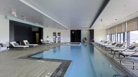 This single-floor indoor aquatic facility offers a refined and tranquil environment centered around a generously sized lap pool with crystal-clear blue water. The pool is framed by smooth tile surrounds and fitted with stainless steel entry railings, ensuring both safety and elegance. Depth markers are clearly visible along the pool's edge, reflecting the space's functional design.

Surrounding the pool, a collection of contemporary lounge chairs and chaise lounges invite relaxation, positioned along both sides of the pool deck. The décor features a neutral palette of whites and grays, complemented by dark accent panels and cabinetry that lend a modern, upscale aesthetic. A distinctive hanging egg chair adds a stylish focal point near the far end of the space.

The ceiling is finished in a smooth, medium-toned gray that adds a sense of enclosure and warmth, while recessed lighting ensures the area is evenly and comfortably illuminated throughout. Large windows along one wall allow natural light to filter in, enhancing the open and airy atmosphere. Private enclosures or changing areas are visible at the rear of the space, adding to the facility's comprehensive amenity offering. Overall, the space exudes a calm, resort-like ambiance suited for both active use and leisure.
