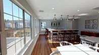This single-floor space offers an open, airy layout distinguished by warm hardwood flooring and soft gray walls that create a clean, contemporary aesthetic. A prominent central feature is the large white bar-style counter, which provides generous seating along both sides via wood-framed bar stools with dark cushioned seats. Above the counter, a row of sleek industrial-style pendant lights in brushed metal adds focused illumination and a modern design element. The walls are decorated with a set of framed botanical artwork, contributing a subtle, refined touch to the space. A generous bank of large double-hung windows lines one side of the room, flooding the interior with abundant natural light and offering views of the surrounding exterior. Recessed ceiling lighting supplements the natural light, ensuring the space remains well-lit throughout. The layout is spacious and versatile, with additional white seating visible in the foreground, suggesting the area is well-suited for communal gathering, dining, or social events. The overall ambiance is bright, welcoming, and thoughtfully appointed with a cohesive blend of modern and transitional design elements.