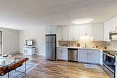 Located at 50 Dinsmore Ave in Framingham, Massachusetts, this well-appointed two-bedroom, two-bathroom residence offers a comfortable and functional single-floor layout. The open-concept kitchen and dining area is a standout feature, showcasing crisp white shaker cabinetry, stainless steel appliances including a refrigerator, dishwasher, microwave, and range, as well as light countertops and hardwood-style flooring that flows throughout the space. The entrance greets you with a painted white brick accent wall, lending a distinctive architectural character to the home. The living room is bright and inviting, with natural light and wall-unit climate control, providing a relaxed atmosphere for everyday living. The spacious primary bedroom offers a window with outdoor views, hardwood-style floors, and ample room for furnishings. The second bedroom serves equally well as a home office or guest room, also featuring a window and natural light. Both bathrooms are finished in a clean, neutral palette with white cabinetry and hardwood-style flooring. With six closets distributed throughout, storage is plentiful. A private balcony extends the living space outdoors, overlooking a landscaped setting with mature trees and a community gazebo.