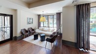 This single-floor communal space is thoughtfully designed to serve multiple functions, combining comfort and productivity in a cohesive, modern aesthetic. Rich hardwood flooring flows seamlessly throughout, anchoring each zone with warmth and continuity.

The lounge area is anchored by a generously sized, dark sectional sofa paired with a rustic wood coffee table and a statement chandelier overhead, creating an inviting gathering space. Adjacent to this, a more intimate seating nook features a white sofa, leather accent chairs, and large floor-to-ceiling windows that flood the area with natural light and offer views of an outdoor pool area.

A dedicated business center is tucked along one wall, featuring a built-in workstation lined with desktop computers beneath a row of stylish pendant lights. A substantial reclaimed wood communal table with curved white chairs serves as a central workspace or meeting area nearby.

A refreshment station with dark cabinetry, counter space, and coffee equipment provides convenient amenities. The overall design balances industrial and contemporary elements, with glass pendant lighting, clean white walls, and dark accents contributing to a polished yet welcoming atmosphere throughout.