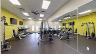 This fitness center is a comprehensively equipped single-floor exercise facility designed to support a wide range of workout routines. The space features a drop-panel ceiling with recessed fluorescent lighting and multiple ceiling fans that promote air circulation throughout the room, contributing to a comfortable workout environment.

The flooring consists of durable dark rubber matting, providing traction and impact absorption suitable for high-intensity activity. A defining architectural feature is the full wall-to-wall mirror installation along one side of the room, which visually expands the space and allows users to monitor their form during exercise.

The cardio zone lines the windowed wall and includes several TRUE-brand treadmills and an elliptical machine. Multiple windows fitted with horizontal blinds allow natural light to filter into this area. The strength training section is centrally and laterally positioned and includes a barbell rack with weight plates, a cable machine, a multi-station power rack, and a tiered dumbbell rack. Spin bikes and additional benches further diversify the training options available.

Bold yellow accent walls add energy to the otherwise neutral-toned interior, while wall-mounted televisions provide entertainment. A glass-panel entry door and emergency exit signage are also present, reinforcing a functional and safety-conscious design throughout.