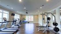 Located at 337 Savannah Hwy in Beaufort, South Carolina, this fitness center is housed in a single-story facility designed to support a well-rounded workout experience. The space features rich hardwood-style flooring that runs throughout, providing a clean and durable surface suited for a variety of exercise activities. Recessed lighting is distributed evenly across the smooth ceiling, complemented by a ceiling fan that promotes air circulation during workouts. Crown molding adds a polished architectural detail, lending the space a finished, upscale feel uncommon in typical gym environments. Large windows along the walls allow generous natural light to fill the room, creating an open and energizing atmosphere. The layout is thoughtfully organized, with a dedicated cardio zone featuring treadmills and elliptical machines positioned near the windows. A functional training area includes stability balls, resistance accessories, and exercise mats. On the opposite side, a weight bench and multi-use strength training rack are positioned alongside wall-mounted mirrors, which visually expand the space and allow for form monitoring. Overall, the fitness center offers a bright, well-appointed environment suitable for a range of fitness routines.