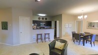This single-floor residence offers an open-concept layout that effortlessly connects the main living, dining, and kitchen areas into one cohesive and functional space. The living area is carpeted and comfortably appointed, anchored by a leather accent chair and ceiling fan overhead, creating a relaxed and inviting atmosphere.

The kitchen is defined by dark cabinetry contrasted against light-toned walls, and is equipped with stainless steel appliances including a microwave. A raised breakfast bar with wooden bar stools serves as both a casual dining spot and a natural visual divider between the kitchen and living areas, making it ideal for entertaining or everyday use.

Adjacent to the kitchen, a dedicated dining area provides additional seating and is framed by framed wall art, adding a refined touch to the space. A pantry or storage closet is conveniently tucked nearby, offering practical organizational solutions.

The space is finished with neutral wall tones and light carpeting in the living zones, transitioning to hard flooring in the kitchen. Subtle décor elements, including decorative plates displayed above the kitchen cabinets, contribute to a warm, homey character throughout. The overall layout is thoughtfully designed to maximize flow and usability across the entire floor.