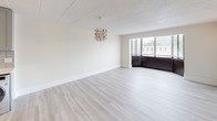 This single-floor unit presents a clean, contemporary aesthetic throughout its thoughtfully arranged layout. The living area is generously proportioned, featuring light-toned hardwood-style flooring and crisp white walls that amplify the sense of openness. A decorative chandelier serves as a focal point, while a large window along one wall invites natural light and provides views of the surrounding neighborhood. Wall sconces add warm accent lighting to complement the airy atmosphere.

The galley-style kitchen is efficiently designed with gray shaker cabinetry lining both sides, offering ample storage from floor to ceiling. White subway tile backsplash, light stone countertops, and a stainless steel sink contribute to the modern, cohesive look. Stainless steel appliances—including a range, over-the-range microwave, and refrigerator—complete the well-equipped cooking space. Light hardwood-style flooring continues seamlessly from the living area into the kitchen.

The bathroom is finished in a bright, neutral palette with clean white surroundings. A full soaking tub with an overhead shower, built-in corner shelving, and chrome fixtures provide a functional and polished bathing space. The overall unit maintains a fresh, move-in-ready presentation with consistent finishes throughout.