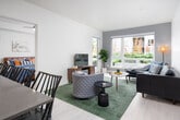 This well-appointed single-floor residence showcases a thoughtfully designed open-concept layout that seamlessly connects the kitchen, dining, and living areas. The kitchen is a standout feature, outfitted with warm wood-toned cabinetry, a subway tile backsplash, dark quartz countertops, stainless steel appliances, and a central island with pendant lighting overhead. Light wood flooring runs throughout the main living spaces, lending the interior a clean, contemporary feel. The living room is bright and welcoming, anchored by oversized windows that frame outdoor greenery and fill the space with natural light. The entry foyer offers a practical transition point between the front door and the main living areas. Two well-sized bedrooms each feature plush carpeting, ceiling fans or lighting fixtures, and windows overlooking the surrounding landscaping. Both bathrooms are finished with dark stone-look countertops, wood-faced vanity cabinetry, undermount sinks, and tub-shower combinations. The overall aesthetic is modern and cohesive, blending neutral tones with warm wood accents to create a comfortable and stylish living environment throughout.