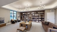 Located at 335 Righters Ferry Rd in Bala Cynwyd, PA, this sophisticated lounge space offers a thoughtfully curated interior designed for comfort and style. The room is anchored by a striking tray ceiling with integrated ambient lighting and complemented by two contemporary globe-and-branch chandeliers that serve as focal points overhead. Recessed lighting throughout further enhances the warm, inviting atmosphere.

A floor-to-ceiling built-in shelving unit spans one wall, displaying an artful arrangement of books, sculptures, and decorative objects that add character and visual depth to the space. The open layout accommodates multiple distinct seating areas: a central grouping featuring a substantial wood block coffee table paired with tufted ottomans and plush sofas, and a cozy L-shaped built-in banquette nestled in a corner, adorned with decorative wall art and accent pillows.

Large floor-to-ceiling windows and multi-pane windows along two sides of the room flood the interior with natural light while offering views of the surrounding exterior. Neutral-toned carpeting and warm taupe walls create a cohesive, calming backdrop throughout. The overall ambiance is modern yet approachable, making this space well-suited for both relaxed socializing and quiet, contemplative use.