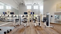 This fitness center in Laurel, Maryland, is organized into two distinct functional areas designed to support a comprehensive workout experience. The main exercise floor features soaring ceilings with coffered detailing, crown molding, and ceiling fans that contribute to an airy, upscale atmosphere. Large upper-level windows flood the space with natural light, while wall-mounted televisions and full-length mirrors line the perimeter, enhancing both entertainment and form-checking during workouts. The equipment selection includes a variety of strength-training machines—notably Life Fitness and Duo-Tek units—alongside treadmills, ellipticals, and a dumbbell rack, providing options for both cardiovascular and resistance training. French doors connect the main gym floor to a separate studio room, which is finished with smooth hardwood-style flooring and floor-to-ceiling mirrors on multiple walls. This open, unobstructed studio space is well-suited for yoga, dance, stretching, or group fitness classes, and is equipped with a wall-mounted television and speaker. Multiple windows throughout the studio invite natural light and views of the surrounding greenery. The overall design balances an elegant architectural aesthetic with practical, modern fitness amenities.