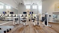 This fitness center in Laurel, Maryland, is organized into two distinct functional areas designed to support a comprehensive workout experience. The main exercise floor features soaring ceilings with coffered detailing, crown molding, and ceiling fans that contribute to an airy, upscale atmosphere. Large upper-level windows flood the space with natural light, while wall-mounted televisions and full-length mirrors line the perimeter, enhancing both entertainment and form-checking during workouts. The equipment selection includes a variety of strength-training machines—notably Life Fitness and Duo-Tek units—alongside treadmills, ellipticals, and a dumbbell rack, providing options for both cardiovascular and resistance training. French doors connect the main gym floor to a separate studio room, which is finished with smooth hardwood-style flooring and floor-to-ceiling mirrors on multiple walls. This open, unobstructed studio space is well-suited for yoga, dance, stretching, or group fitness classes, and is equipped with a wall-mounted television and speaker. Multiple windows throughout the studio invite natural light and views of the surrounding greenery. The overall design balances an elegant architectural aesthetic with practical, modern fitness amenities.